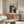 May include: Black and white abstract art poster with paint splatters and drips displayed above a mid-century modern walnut credenza. Room features cream walls, white sculptural chair, glass oval coffee table, and warm wood tones throughout. Poster shows dramatic contrast with organic paint patterns against light background in thin frame.