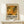 May include: Framed exhibition poster with abstract artwork in warm oranges, yellows and greens displayed on light wood console table. Poster features bold black text and dates, housed in natural wood frame. Small white wrapped gift with greenery sits beside frame against cream colored wall.