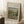 May include: Framed Renoir landscape painting print showing lush green chestnut trees in an impressionist style, displayed in a light wood frame against white wall corner. The artwork features soft brushstrokes in greens, blues and earth tones depicting a pastoral scene. Scene includes white coffee cup on wooden block and beige textured throw blanket nearby, creating a cozy home interior setting with natural lighting.