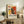 May include: Abstract art poster with vibrant red, yellow, and black paint splatters in Pollock style, displayed in light wooden frame on rustic sideboard. Room setting features fern plant, cream ceramic vase, stacked books, and brown leather chair. Poster shows chaotic brushstrokes and drip painting technique with exhibition text below artwork.