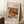May include: Framed art print leaning against white wall on textured white rug, featuring Renoir's still life painting of colorful fruit including red apples, green apple, and citrus fruits. Light wood frame with museum-style white matting and black text. Coffee cup on wooden block visible to left, beige fabric draped on right. Natural window light creates soft shadows across the scene.