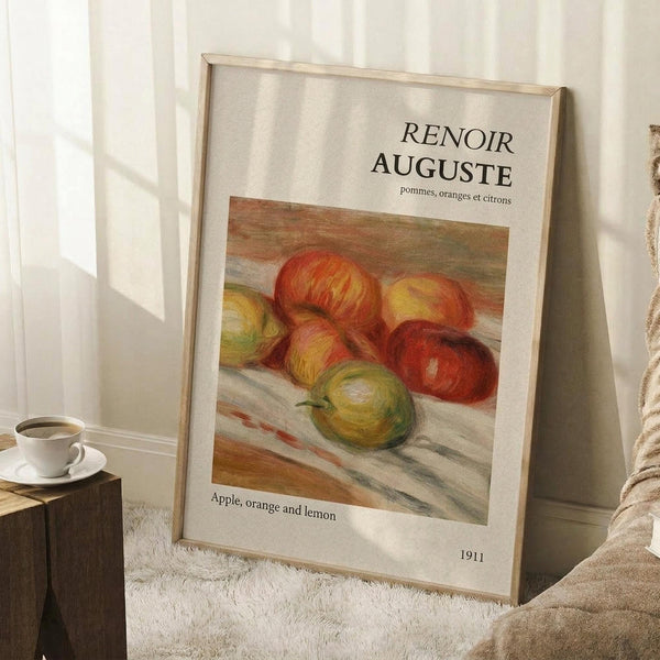 May include: Framed art print leaning against white wall on textured white rug, featuring Renoir's still life painting of colorful fruit including red apples, green apple, and citrus fruits. Light wood frame with museum-style white matting and black text. Coffee cup on wooden block visible to left, beige fabric draped on right. Natural window light creates soft shadows across the scene.