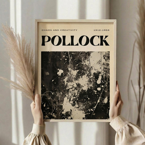 May include: Hands holding a framed poster featuring Jackson Pollock's abstract expressionist drip painting in black and white, with text reading "Chaos and Creativity" and "1912-1956" at top. Light wood frame against neutral beige clothing and dried pampas grass in soft natural lighting.
