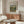 May include: Vintage Renoir landscape art print in light wooden frame hanging above mid-century walnut credenza in minimalist living room. Print shows impressionist painting of chestnut trees in warm greens and earth tones. Room features cream textured chair, glass-top oval coffee table, modern table lamp, and soft natural lighting on white walls.