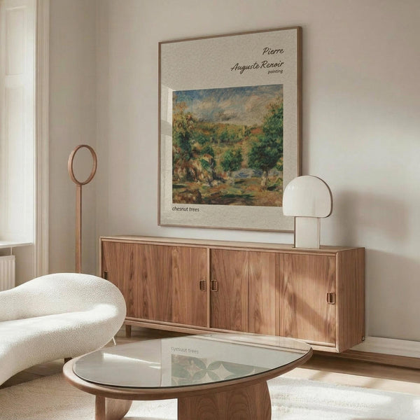 May include: Vintage Renoir landscape art print in light wooden frame hanging above mid-century walnut credenza in minimalist living room. Print shows impressionist painting of chestnut trees in warm greens and earth tones. Room features cream textured chair, glass-top oval coffee table, modern table lamp, and soft natural lighting on white walls.