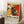 May include: Abstract expressionist style poster in light wooden frame leaning against white wall corner. Features dynamic splashes and drips in red, yellow, black and white paint with bold brushstrokes and splatters. Exhibition-style design with "POLLOCK" and "EMBRACE THE CHAOTIC REALM OF CREATIVITY" text at bottom. Scene shows cozy room setting with white coffee cup on wooden block side table and cream knit throw blanket on white textured rug.