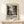 May include: Framed poster with black and white abstract splatter artwork displayed on light wood shelf against cream wall. Wood frame surrounds vintage-style exhibition poster featuring chaotic paint splatters and drips. Small wrapped gift box with greenery sits beside frame. Natural lighting creates soft shadows on minimal modern interior setting.