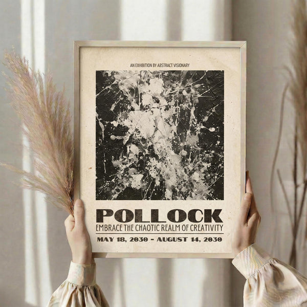 May include: Person holding a framed vintage-style poster featuring black and white abstract splatter artwork reminiscent of Jackson Pollock's style, with exhibition text reading "AN EXHIBITION BY ABSTRACT VISIONARY" and "POLLOCK EMBRACE THE CHAOTIC REALM OF CREATIVITY" with dates "MAY 18, 2030 - AUGUST 14, 2030". Light wood frame against cream matting, held alongside dried pampas grass in soft natural lighting near a window.