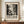 May include: Framed vintage-style exhibition poster with black and white abstract splatter artwork resembling Jackson Pollock's drip painting technique. Features bold "POLLOCK" text and exhibition dates from May to August 2030. Poster displayed on wooden sideboard next to white ceramic vase and stacked books in elegant room with cream walls and white wainscoting. Brown wooden frame complements the warm wood furniture tones.