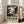 May include: Large framed abstract art poster with black paint splatters and drips on cream background, featuring bold "POLLOCK" text and exhibition details. Poster leans against white wall next to beige upholstered chair in bright living room with tall windows and natural lighting.