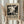 May include: Person holding framed black and white abstract art poster with paint splatter design reminiscent of Jackson Pollock's drip painting style. Beige wooden frame against neutral cream curtained background. Dried pampas grass visible on left side. Poster features bold text reading "POLLOCK" and "EMBRACE THE CHAOTIC REALM OF CREATIVITY" with exhibition dates. Person wearing cream ribbed sweater with gathered cuffs.