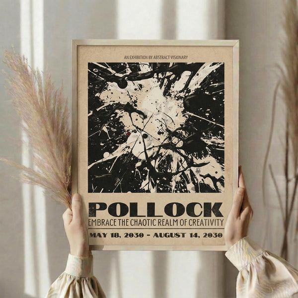 May include: Person holding framed black and white abstract art poster with paint splatter design reminiscent of Jackson Pollock's drip painting style. Beige wooden frame against neutral cream curtained background. Dried pampas grass visible on left side. Poster features bold text reading "POLLOCK" and "EMBRACE THE CHAOTIC REALM OF CREATIVITY" with exhibition dates. Person wearing cream ribbed sweater with gathered cuffs.
