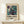 May include: Framed abstract art poster with swirling teal, purple, and cream marbled patterns displayed on light wooden shelf against white wall. Features bold "POLLOCK" text and exhibition-style layout with dates. Small wrapped gift box with greenery sits beside the natural wood frame.