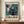 May include: Framed abstract art poster displayed on wooden sideboard next to white ceramic vase and stacked books. The artwork features swirling marbled patterns in teal, purple, and cream colors with fluid, organic shapes. Poster has cream matting and dark wood frame, shown against pale wall with white trim molding in elegant interior setting.