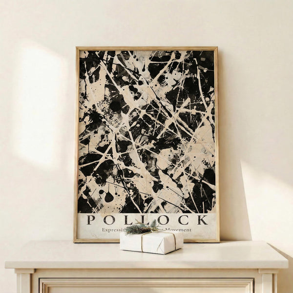 May include: Black and white abstract expressionist art print displayed in light wood frame on cream wooden surface against beige wall. Artwork features dynamic paint splatters, drips, and gestural marks in monochromatic palette. Small wrapped gift box with evergreen sprig sits beside frame. Print shows bold contrast between dark paint strokes and light background with energetic, spontaneous brushwork typical of action painting style.