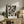 May include: Black and white abstract expressionist artwork with dynamic paint splatters and gestural brushstrokes in wooden frame, displayed on rustic wood sideboard alongside table lamp with cream shade, potted fern, stacked books, and ceramic vase in neutral-toned living room setting