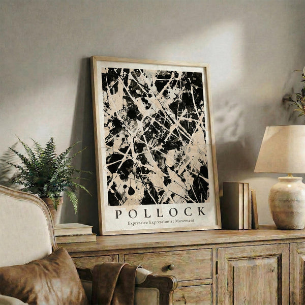 May include: Black and white abstract expressionist artwork with dynamic paint splatters and gestural brushstrokes in wooden frame, displayed on rustic wood sideboard alongside table lamp with cream shade, potted fern, stacked books, and ceramic vase in neutral-toned living room setting