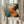 May include: Person holding framed abstract expressionist art print featuring dynamic paint splatters and brushstrokes in turquoise, orange, red, and black colors. Light wooden frame against neutral curtained background with dried pampas grass visible to the side. Bold gestural movements create energetic composition with paint drips and textural elements throughout the square format artwork.