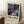 May include: Framed abstract art poster with cosmic swirling patterns in deep purple, teal, and navy blue colors, accented with golden dots and flowing lines. The poster features exhibition-style text layout with dates, displayed on a white fluffy rug beside a wooden side table holding a white coffee cup and books. Natural window light creates soft shadows on white walls.