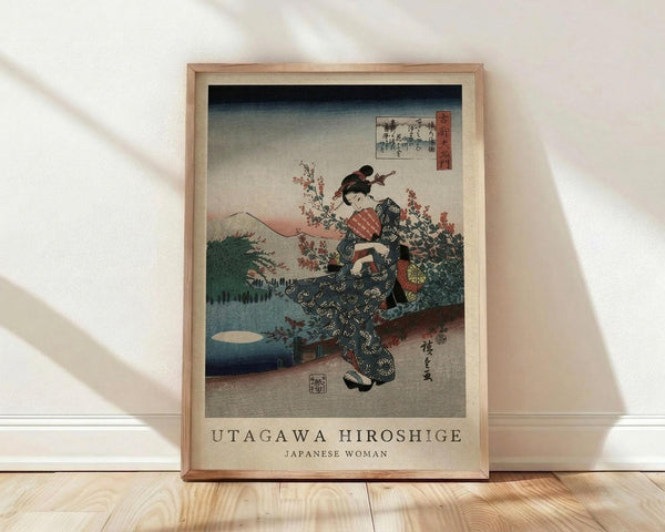 May include: Framed vintage Japanese woodblock print showing a woman in ornate dark kimono with floral patterns standing among cherry blossoms. Mount Fuji visible in background with gradient sky from dark blue to pale yellow. Print has traditional Japanese text and red seal marks. Natural wood frame sits on light wooden floor against white wall with soft diagonal shadows.