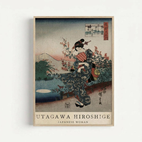 Japanese woodblock print depicting a woman in an elaborate dark kimono with circular patterns, kneeling among cherry blossoms. The scene includes Mount Fuji in the background, traditional Japanese calligraphy, and red seal stamps. The artwork features muted colors with blue, pink, and earth tones in a classic ukiyo-e style. Displayed in a light wooden frame against a white wall.
