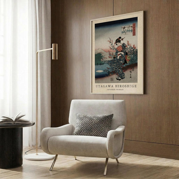 May include: A framed vintage Japanese woodblock print displayed on a wood-paneled wall above a beige upholstered armchair with geometric pillow. The artwork shows a woman in colorful traditional dress with floral elements against a blue and cream background. Modern brass floor lamp and dark round side table with open book complete the neutral-toned room scene.