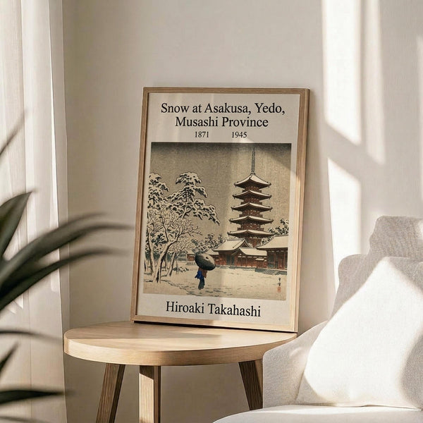 May include: Framed vintage Japanese woodblock print showing snow-covered pagoda temple with figure holding umbrella, displayed on round wooden side table next to beige upholstered chair. Soft natural lighting creates shadows on white wall behind frame. Green plant leaves visible in corner. Print features muted earth tones with traditional Japanese winter scene architecture and snowy trees.