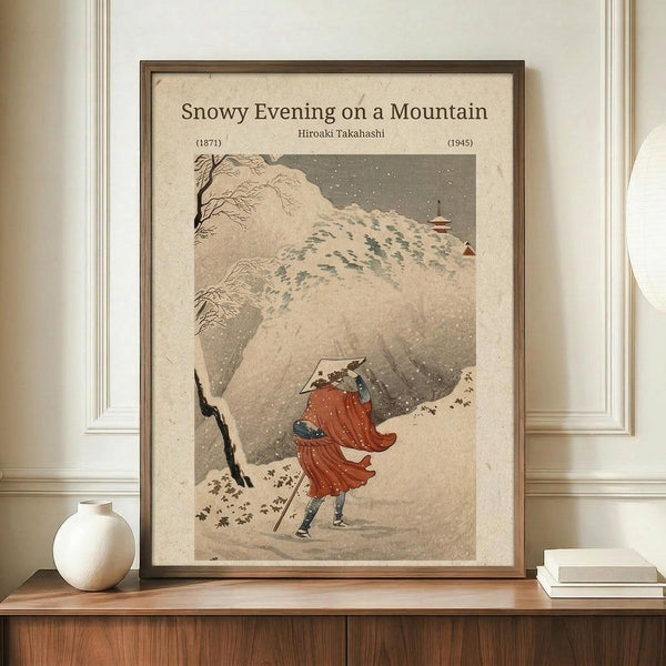 May include: Japanese woodblock-style poster showing a figure in flowing red robes walking through a snowy mountain landscape with falling snow and bare trees. The artwork is displayed in a brown wooden frame on a wooden sideboard next to a white ceramic vase and books, against cream colored walls with white trim molding.