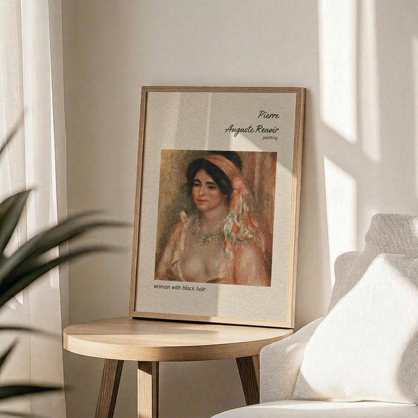 May include: Framed art print featuring a Renoir painting of a woman with dark hair wearing an orange headband and flowing white dress, displayed on a light wooden round side table next to a beige upholstered chair. Natural sunlight creates soft shadows across the neutral-toned room setting. Green plant leaves visible in corner, creating a warm, minimalist home decor scene.