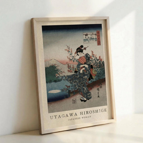 May include: Framed vintage Japanese woodblock print reproduction showing a woman in traditional kimono with floral patterns standing among cherry blossoms. Mountains and water visible in background with gradient sky from blue to pink. Light wooden frame displayed against white wall on light surface. Print features classic ukiyo-e art style with rich colors and delicate details.