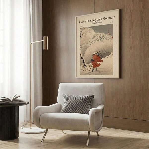 May include: Framed vintage Japanese woodblock print showing figure in red clothing walking through snowy mountain landscape with walking stick. Displayed on wood paneled wall above beige upholstered armchair with geometric patterned pillow. Modern reading corner with brass floor lamp and dark side table holding open book.
