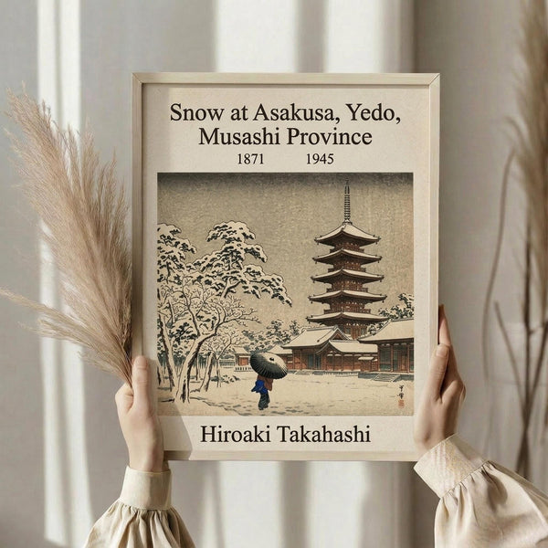 May include: Person in cream sweater holding framed vintage Japanese woodblock print showing snowy temple scene with pagoda and figure with umbrella. Light wooden frame against white curtained background with dried pampas grass decoration. Muted earth tones and traditional ukiyo-e art style visible in sepia-toned print.