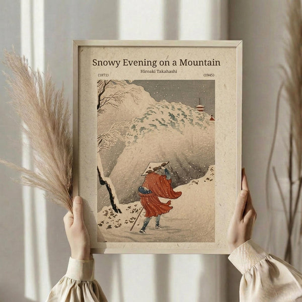 May include: Hands holding a framed vintage Japanese woodblock print showing a figure in red robes walking through falling snow on a mountain path, with bare trees and traditional buildings visible in the muted beige and gray winter landscape. Light wooden frame against soft neutral background with dried pampas grass decoration.