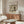 May include: Vintage Japanese woodblock print poster displayed above modern walnut credenza in minimalist living room. Print shows snow-covered temple pagoda with figure holding umbrella, rendered in muted earth tones. Room features cream upholstered chair, glass coffee table, and warm wood furnishings against white walls.