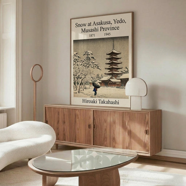 May include: Vintage Japanese woodblock print poster displayed above modern walnut credenza in minimalist living room. Print shows snow-covered temple pagoda with figure holding umbrella, rendered in muted earth tones. Room features cream upholstered chair, glass coffee table, and warm wood furnishings against white walls.