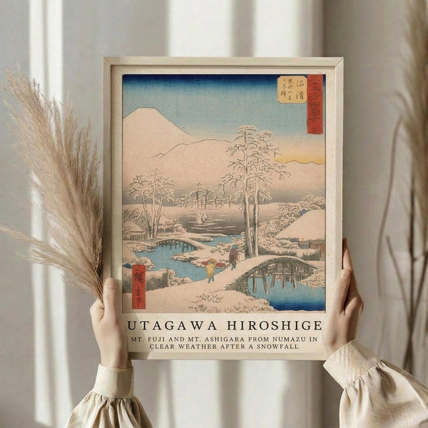 May include: Hands holding a framed reproduction of a Japanese woodblock print showing a snowy winter landscape with Mount Fuji, bare trees, a blue stream, small figures crossing a bridge, and traditional buildings. The artwork features soft blues, whites, and peach tones typical of ukiyo-e style. Light wood frame against cream curtains with dried pampas grass visible on the left side.