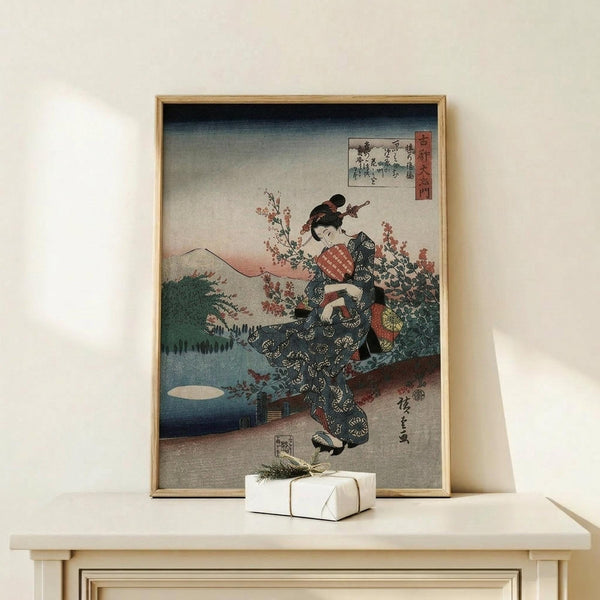 May include: Framed Japanese woodblock print showing a woman in dark kimono with red obi sash among cherry blossoms, with Mount Fuji and lake in background. Print displays in light wood frame on pale wooden surface beside white wrapped gift with twine. Vintage ukiyo-e art style with traditional Japanese text cartouches in upper corners.