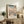 May include: Framed Japanese woodblock print featuring Mount Fuji in winter with snow-covered landscape, bare trees, blue stream, and traditional buildings under gradient sky from blue to peach. Print displays on rustic wooden sideboard alongside fern plant, ceramic vase, and books in neutral-toned living room setting.