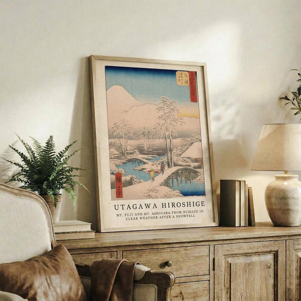 May include: Framed Japanese woodblock print featuring Mount Fuji in winter with snow-covered landscape, bare trees, blue stream, and traditional buildings under gradient sky from blue to peach. Print displays on rustic wooden sideboard alongside fern plant, ceramic vase, and books in neutral-toned living room setting.