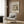 May include: Framed vintage Japanese woodblock print showing snow-covered temple pagoda with figure holding umbrella, displayed on warm wood-paneled wall above beige upholstered armchair with geometric patterned pillow. Modern brass floor lamp and dark round side table with open book complete the cozy reading nook scene with white curtains and light wooden flooring.