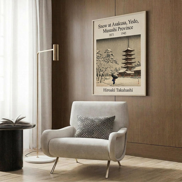 May include: Framed vintage Japanese woodblock print showing snow-covered temple pagoda with figure holding umbrella, displayed on warm wood-paneled wall above beige upholstered armchair with geometric patterned pillow. Modern brass floor lamp and dark round side table with open book complete the cozy reading nook scene with white curtains and light wooden flooring.