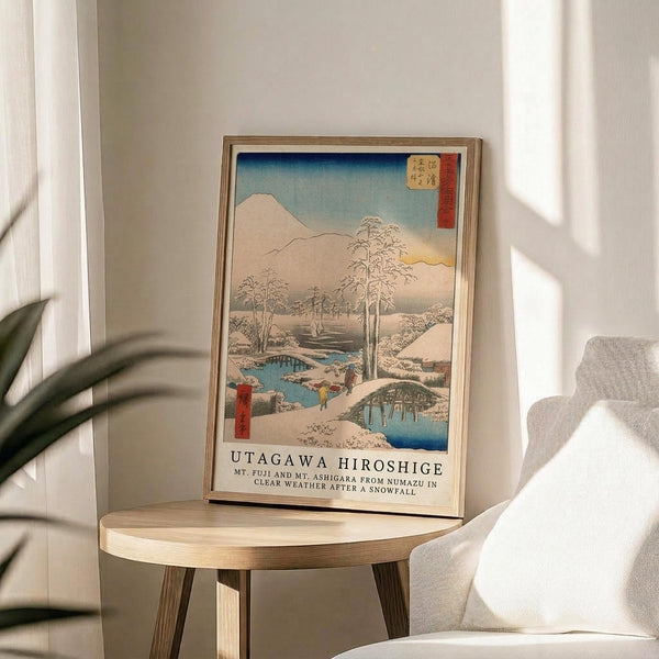 May include: Framed Japanese woodblock print showing snow-covered winter landscape with Mount Fuji, bare trees, blue stream, and small figures crossing a bridge. Print features soft blues, whites, and earth tones with traditional red text cartouche. Wooden frame sits on round light wood side table next to beige upholstered chair and potted plant, with white curtains and natural sunlight streaming across neutral room.