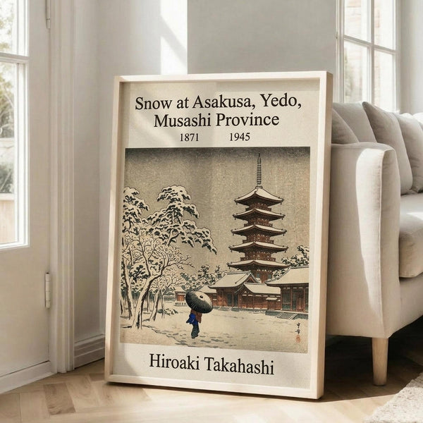 May include: Framed vintage Japanese woodblock print showing snow-covered temple with five-story pagoda and figure with umbrella, displayed in light wooden frame leaning against white wall next to beige upholstered chair in bright modern living room with large windows