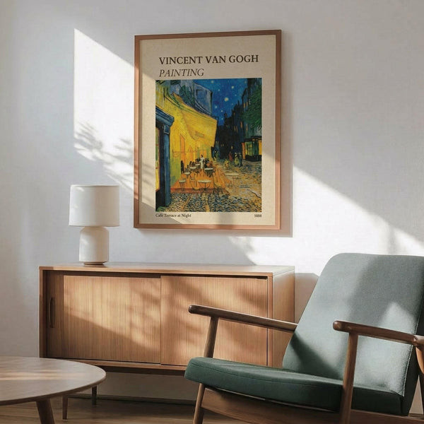 May include: Framed Van Gogh Cafe Terrace at Night poster print displayed above a wooden sideboard in modern living room. Print shows yellow cafe terrace with outdoor seating under starry blue night sky, housed in thin brown frame. Scene includes mint green mid-century armchair with wooden arms, white table lamp, and round coffee table. Warm natural lighting creates shadows on white wall.