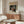 May include: Japanese ukiyo-e woodblock print artwork displayed above a mid-century modern wooden sideboard in a minimalist living room. The colorful print features a warrior figure in traditional dress against a dark snowy background with Japanese text. Scene includes a white curved chair, glass-top oval coffee table, and modern floor lamp with warm lighting on white walls.
