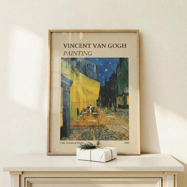 May include: Framed reproduction of Van Gogh's Cafe Terrace at Night painting showing yellow cafe with outdoor seating under starry blue sky on cobblestone street. Print features cream matting in light wood frame, displayed on pale wooden surface alongside small white wrapped gift with greenery. Soft natural lighting creates gentle shadows on neutral wall background.