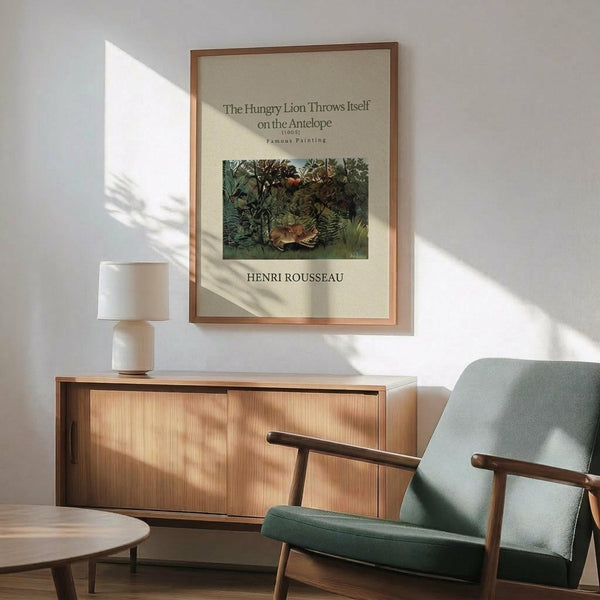 May include: Framed art print of Henri Rousseau's jungle painting featuring a lion attacking an antelope among lush green foliage, displayed above a wooden sideboard with white ceramic lamp in a modern living room with sage green upholstered chair and natural lighting casting shadows on white wall.
