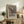 May include: Black and white reproduction of Munch's famous expressionist artwork showing a figure with hands to face in anguish on a bridge, displayed in light wood frame on rustic wooden sideboard. Room features neutral walls, fern plant, ceramic vase, stacked books, and beige upholstered chair with leather cushion. Poster includes bold text identifying the artwork as a masterpiece of expressionism.