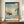 May include: Japanese woodblock print poster featuring a Western sailing ship with multiple masts and colorful flags on choppy blue-green waves, with rocky coastline and pine tree visible on right side. Japanese text cartouche in upper right corner. Print displayed in brown frame on wooden credenza next to white ceramic vase and stacked books in neutral-toned room with white walls and molding.