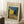 May include: Framed Van Gogh reproduction print showing Cafe Terrace at Night painting with yellow cafe, blue starry sky, and cobblestone street. Light wood frame with cream matting, leaning against white wall corner. Small wooden side table with white coffee cup and saucer nearby. Soft natural lighting creates gentle shadows on white fluffy area rug.